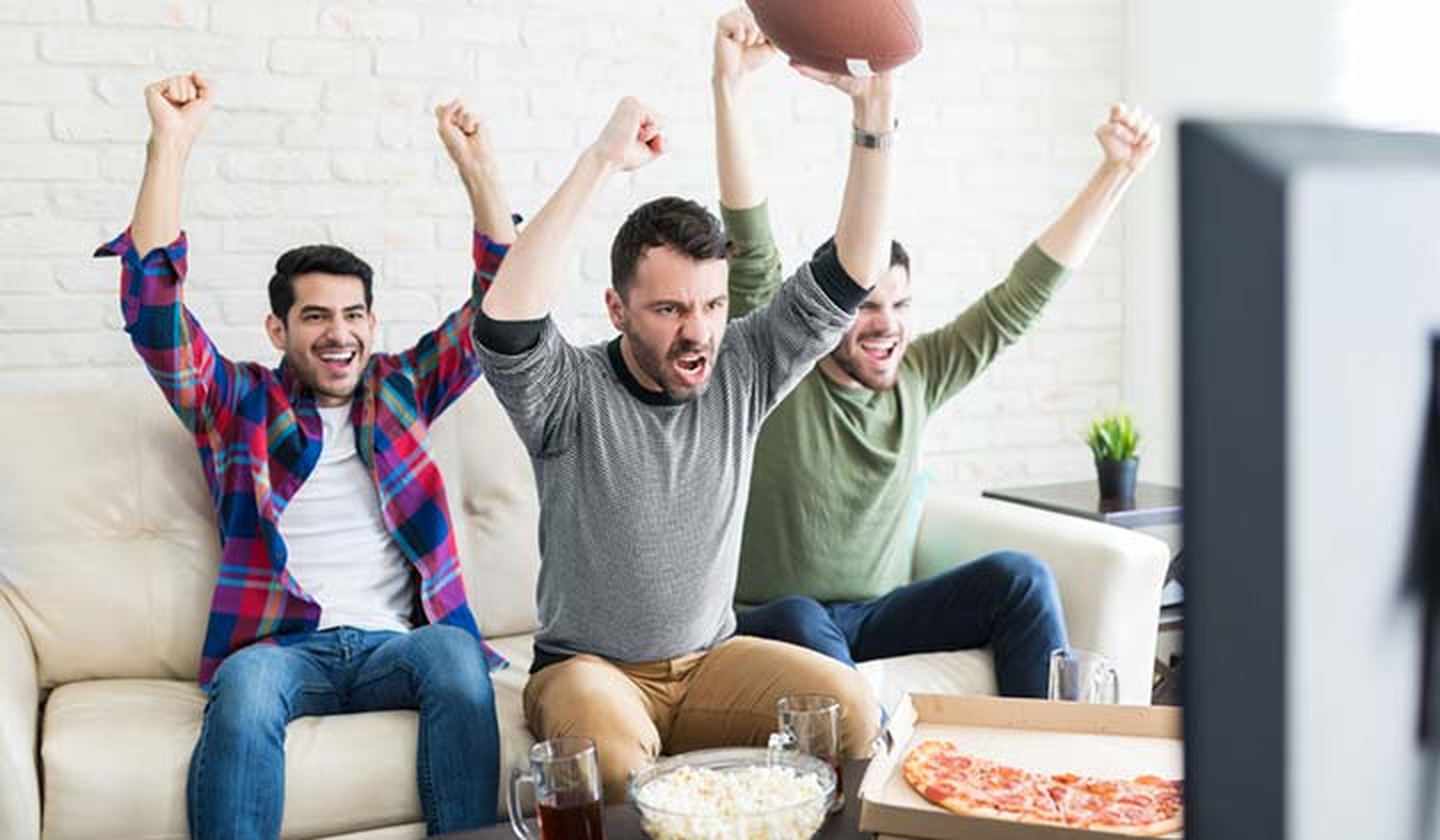 Young men holding footballs cheering while watching TV