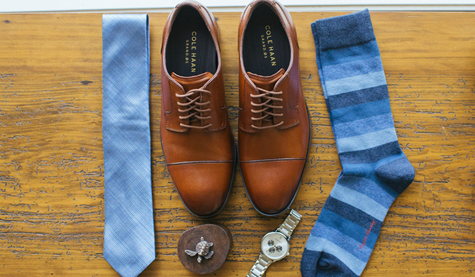 Cole Haan shoes sitting by a blue tie, blue striped socks, and a watch