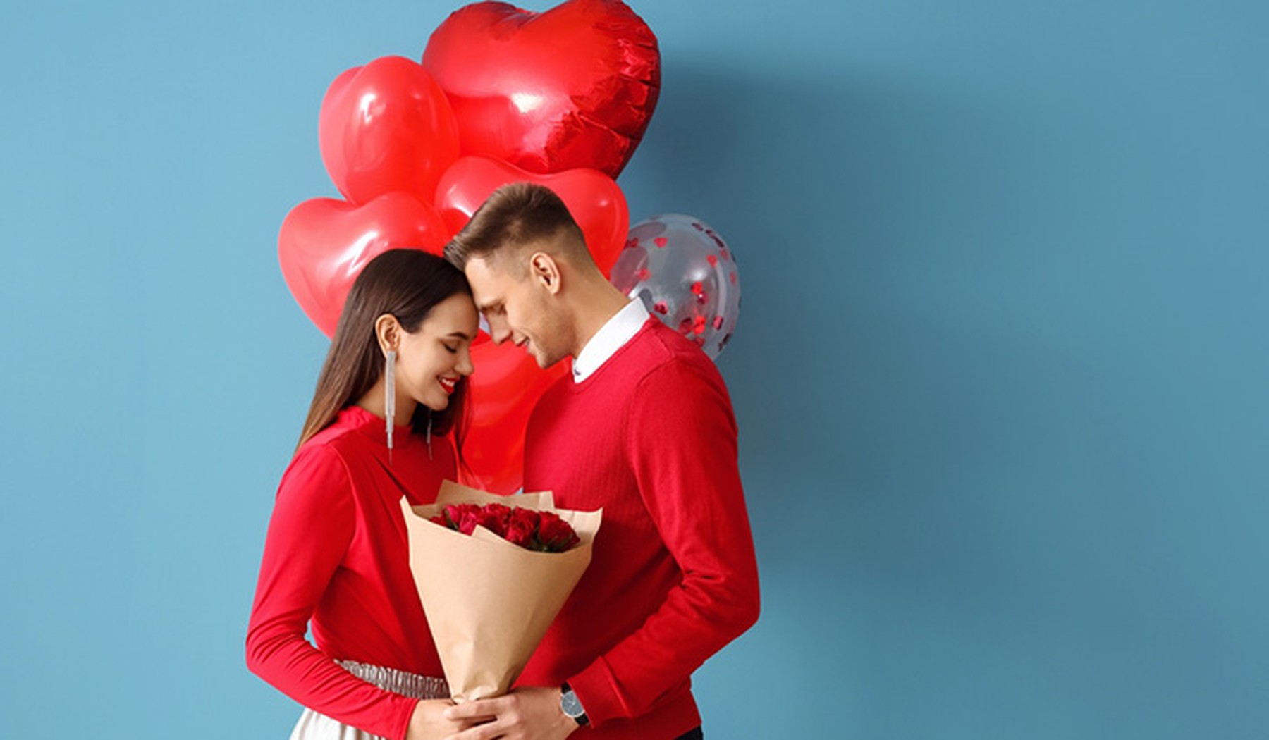 Young man and woman dressed in red in front of heart shaped balloons, holding red roses