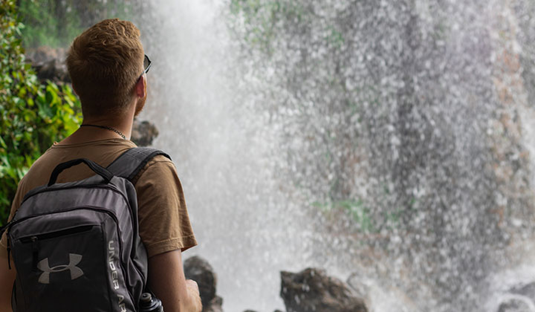 Young man in front of a waterfall with an Under Armour backpack